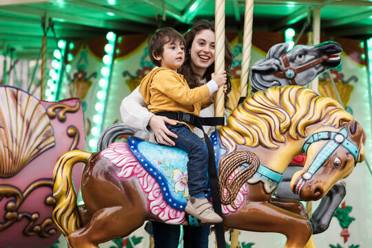 A mother supports her son on a carousel horse at an amusement park