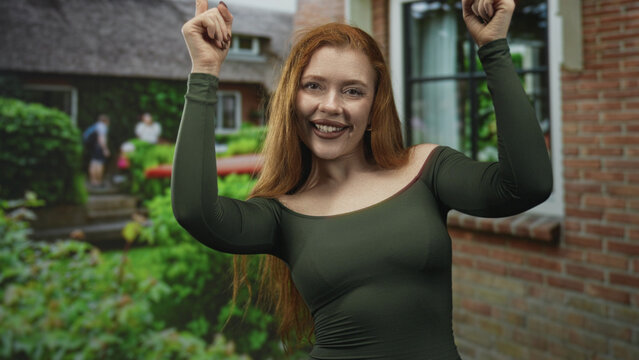 Redhead woman pointing both index fingers upward with bare shoulders visible, smiling in front of a brick building and lush garden; joy celebration.