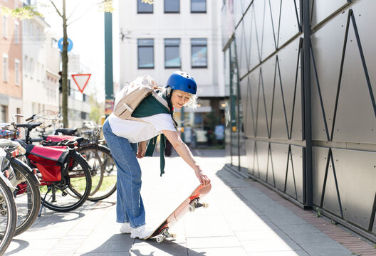 A woman picks up her skateboard on a sunny urban street