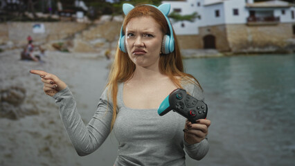 Young woman wearing cat ear headphones holds gamepad and points finger in front of seaside building while pouting and squinting in displeasure  frustration defiance. © Krakenimages.com