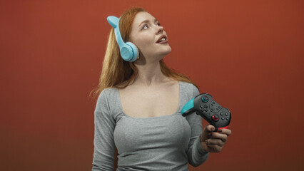 Woman holding a gamepad and wearing blue cat ear headphones, hand gripping a turquoise accented controller against a red studio wall  playful joy. © Krakenimages.com
