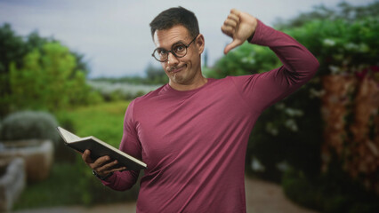 Obraz premium Man with moustache and glasses reading a book while showing a thumbs down gesture in a forest setting with shrubs and a path; disapproval.