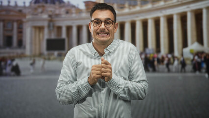 Man clasping hands and grimacing with a nervous smile, fingers interlaced visible, standing before a grand building colonnade and crowd  anxiety awkwardness. © Krakenimages.com