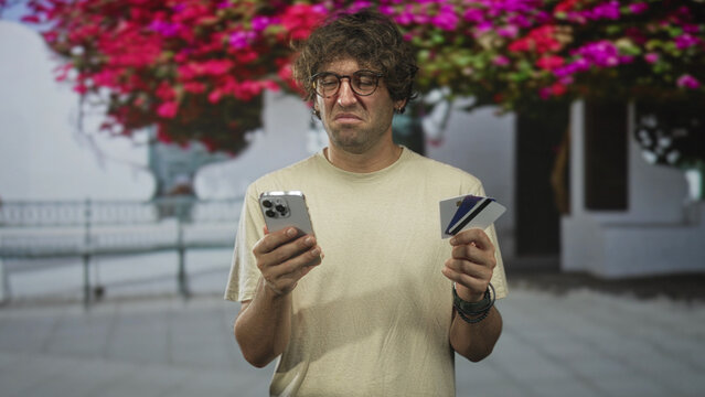 Man holds smartphone and creditcard, grimaces while checking phone in front of building; frustration payment issue.