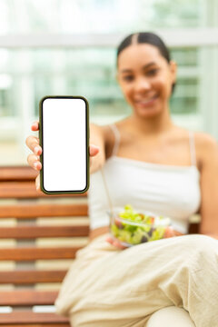 Smiling woman shows a blank smartphone screen while eating a salad