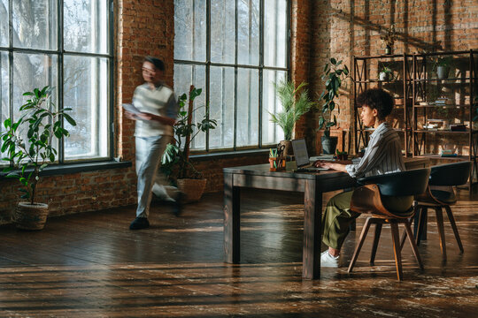 Businesswoman working at laptop while colleague walks in modern office