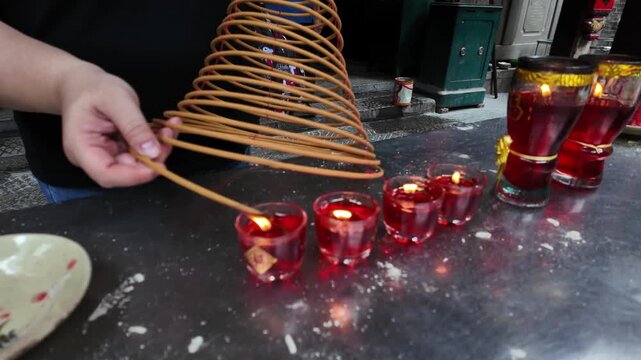 Close-up of a person lighting incense coil from red flame lamps inside a historic Chinese temple in Cholon, Ho Chi Minh City, Vietnam