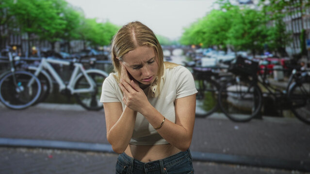 Woman with headphones pressing her neck and wincing while standing on a street beside parked bicycles and a canal; pain fatigue.