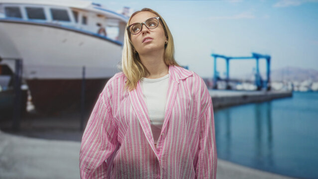 Woman wearing glasses and a pink striped overshirt tilts her head with an unimpressed expression beside a boat in a studio; boredom solitude.