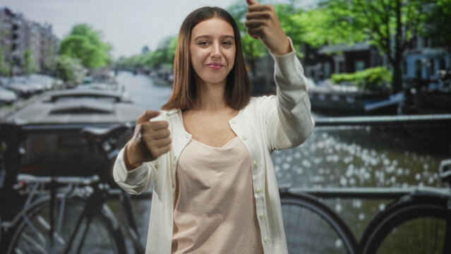 Woman with palm forward and clenched fist miming steering on a street by a canal and bicycle bridge; playful curiosity.