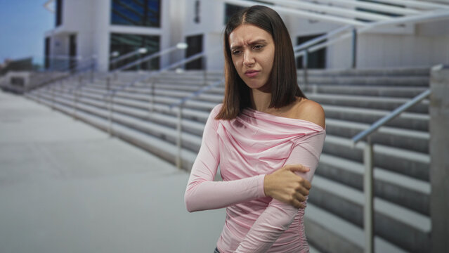 Young woman holds shoulder with hand on concrete building steps outside a modern public building, one shoulder exposed and grimacing; pain.