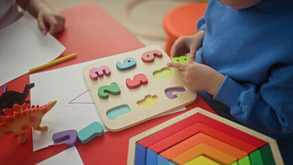 Fototapeta premium Boy placing wooden number puzzle pieces with hands on red table in preschool, colorful rainbow stack, toy dinosaurs, pencil and drawing paper visible; concentration learning playtime.