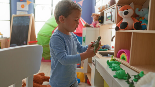 Boy holding green toy dinosaur with bare hands while arranging plastic frogs on a low shelf in a playroom building; curiosity learning.