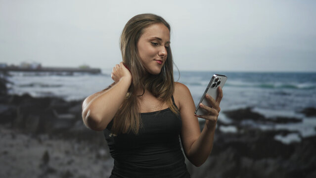Woman holding smartphone and tucking hair on a rocky beach while smiling and checking messages on her phone; serene confidence.