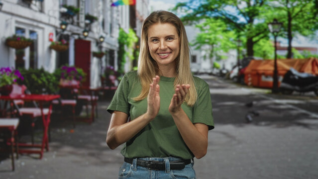 Blonde woman clapping hands on a sunny street beside outdoor cafe tables and parked scooters; celebration joy.
