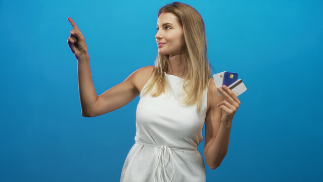 Woman in white dress points finger while holding multiple creditcards against blue studio backdrop; confidence payment choice.