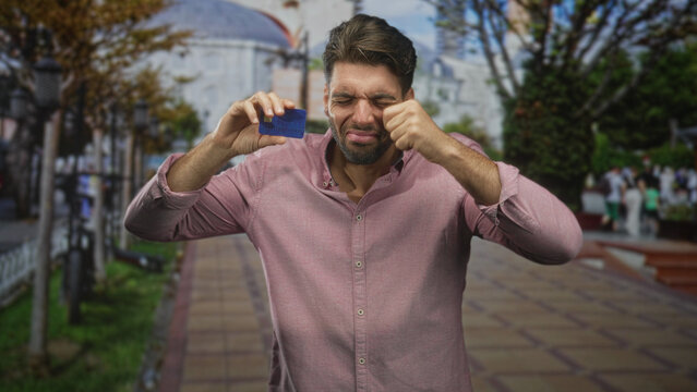 Hispanic man with beard holds a blue creditcard and rubs eye on a street while showing a frustrated payment gesture; payment frustration.