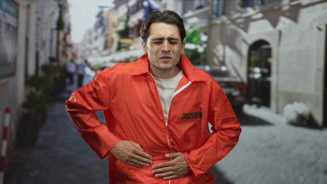 Man in orange jumpsuit clutching his stomach with hands over zipper and inmate number visible on a street lined with bollards and plants; suffering pain.