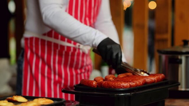 Grilled sausages cooking on electric grill at outdoor food market stall. Street food vendor preparing hot dogs on wooden table. Fast food preparation concept.