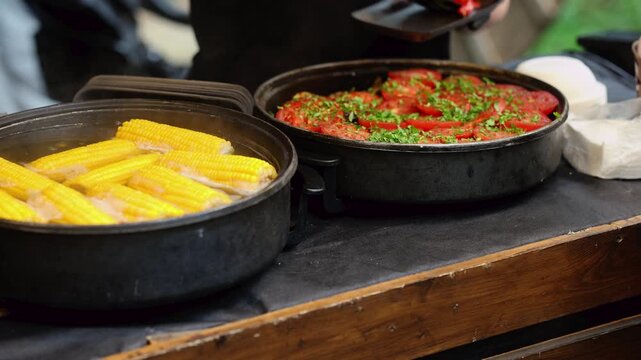 Pots with boiled corn cobs and baked tomatoes with herbs on wooden counter. Traditional street food cooking at outdoor market. Rustic festival cuisine preparation concept