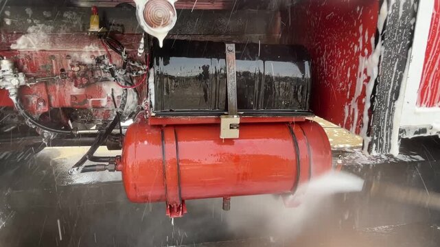Pressure water washes the lower rear section of a red truck, rinsing a cylindrical tank and nearby components while removing dirt from the vehicle underbody. during routine vehicle cleaning.