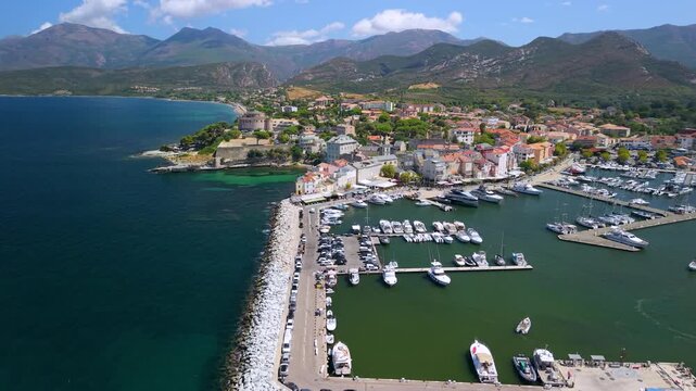 Saint-Florent Corsica island drone marina old town yacht harbor pastel houses turquoise bay. Aerial flyover along pier with moored yachts, panning reveal of city landscape surrounded by mountains 4K.