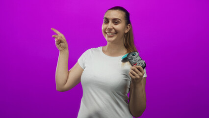 Woman holding a gamepad points finger to the side while smiling, wearing a white t shirt in studio  playful confidence. © Krakenimages.com