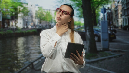 Woman holding tablet touches neck on street by canal while squinting with red glasses  reflective discomfort. © Krakenimages.com
