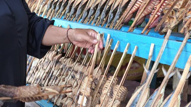 Close-up of a street vendor's hands arranging smoked seafood on bamboo skewers across a vibrant blue wooden shelf in a traditional Asian roadside market.
