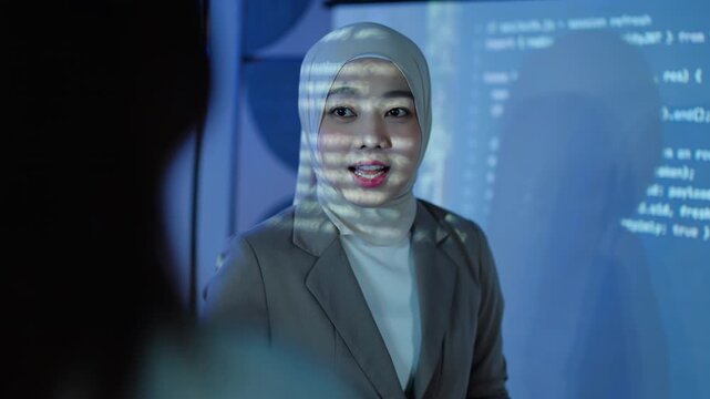 A professional Muslim female software developer wearing a hijab presents program code projected on a screen in a dark office, explaining data analytics and tech solutions to her team at night.