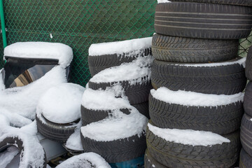 Large piles of used car tires covered with fresh snow stand beside a fenced workshop area. Automotive stock image for winter tire change, tire storage, recycling, vehicle maintenance, rubber disposal, © Volha