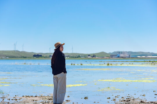 One Asian woman enjoying scenic landscape of salt lake wetland and distant wind turbines in summer in Ulan Hada Volcano Geopark, Ulanqab, Inner Mongolia, China