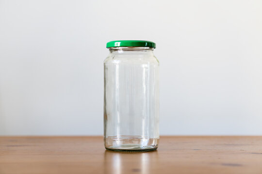 Front view of empty clear glass jar with green metal lid on wooden table against minimalist light grey background with copy space.