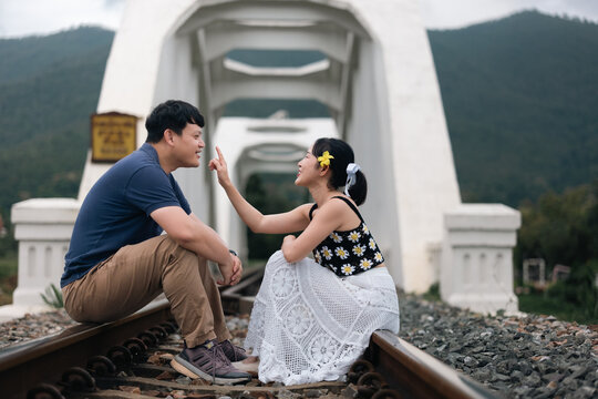 Playful Asian couple sitting on railway tracks in front of historic white bridge