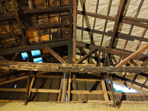 A detailed close-up of a damaged, old wooden roof structure with charred beams and exposed tiles, looking up towards patches of blue sky, showing fire damage.