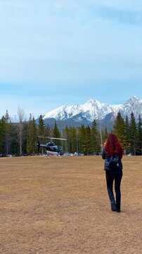 Following a woman with a backpack walking towards the helicopter standing nearby. Pine-tree forest and gorgeous snow-capped mountains in the backdrop. Vertical video.