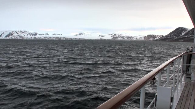 A ship crosses the Arctic Ocean beside snow-covered mountain peaks, showing the scale of the remote northern coast and cold open water. The source identifies the location near Norway.