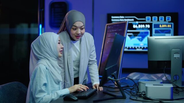 Two diverse Muslim women in hijabs celebrate success while programming in a high-tech office. Female IT developers smile and high five after solving a complex coding problem on computer screens.