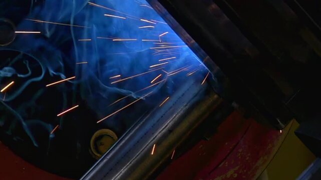 A robotic argon welding arm joins metal pipe sections with controlled motion during an industrial welding process. Bright sparks flare around the welding area as the process continues.