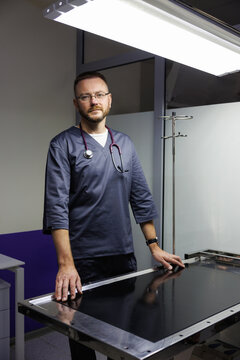 Portrait of doctor standing by examining table and looking at camera