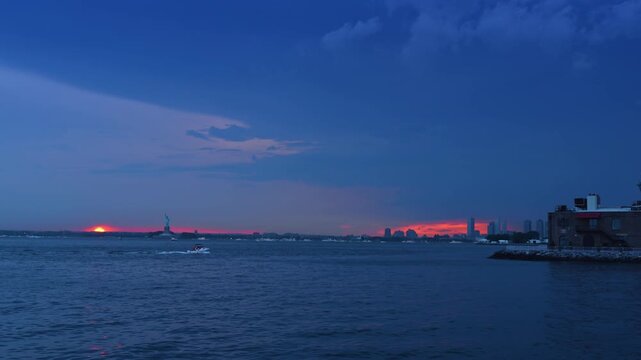 Small motorboat moves by the waterscape at dusk time in New York Harbor. The Statue of Liberty and setting sun in the backdrop.