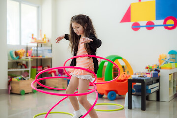 Energetic young Asian girl having fun spinning pink hula hoops in a colorful kindergarten playroom, enhancing physical education, motor skills, and an active healthy childhood development. © VStudio