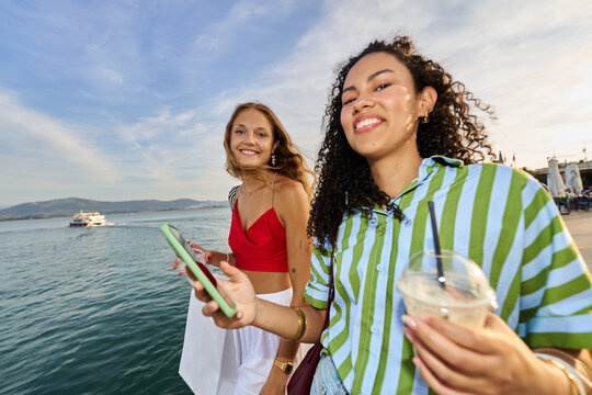 Happy friends enjoying waterfront stroll with drinks