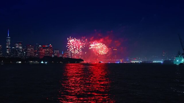 Fireworks ignite over New York skyline with One World Trade Center. Red sparks bloom above water reflecting vibrant glow near downtown.