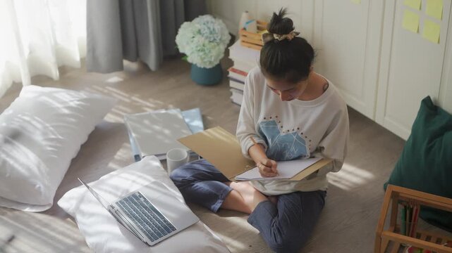 Woman working on laptop sitting on floor by bed, focused studying with sticky notes and books, cozy remote work from home routine in bright bedroom concept