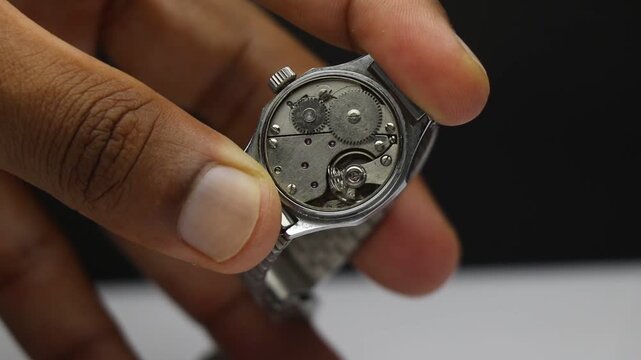 Mechanical watch movement with damaged spring held in fingers showing exposed gears and internal parts needing repair