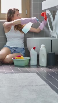 Happy smiling young woman spraying detergent and wiping toilet seat with microfiber cloth