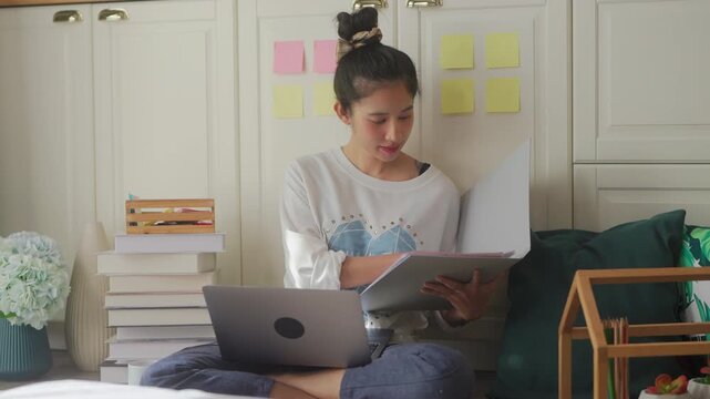Woman working on laptop sitting on floor by bed, focused studying with sticky notes and books, cozy remote work from home routine in bright bedroom concept