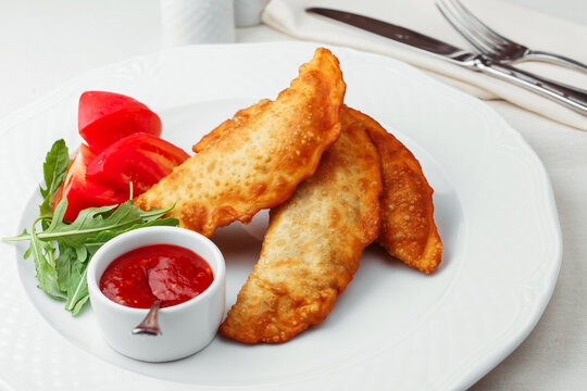 Crispy homemade chebureki with meat filling, tomato dipping sauce, tomatoes and arugula on elegant white plate