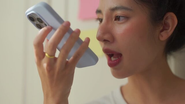A woman speaks into her smartphone using a voice command feature, sitting comfortably in a warm, softly lit kitchen nook surrounded by books and cozy home decor, blending everyday life with smart tech